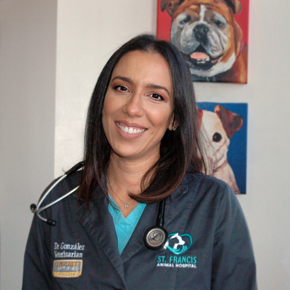 Smiling veterinarian in dark colored scrubs with St Francis Animal Hospital logo wearing a stethoscope. Paintings of dogs in the background.