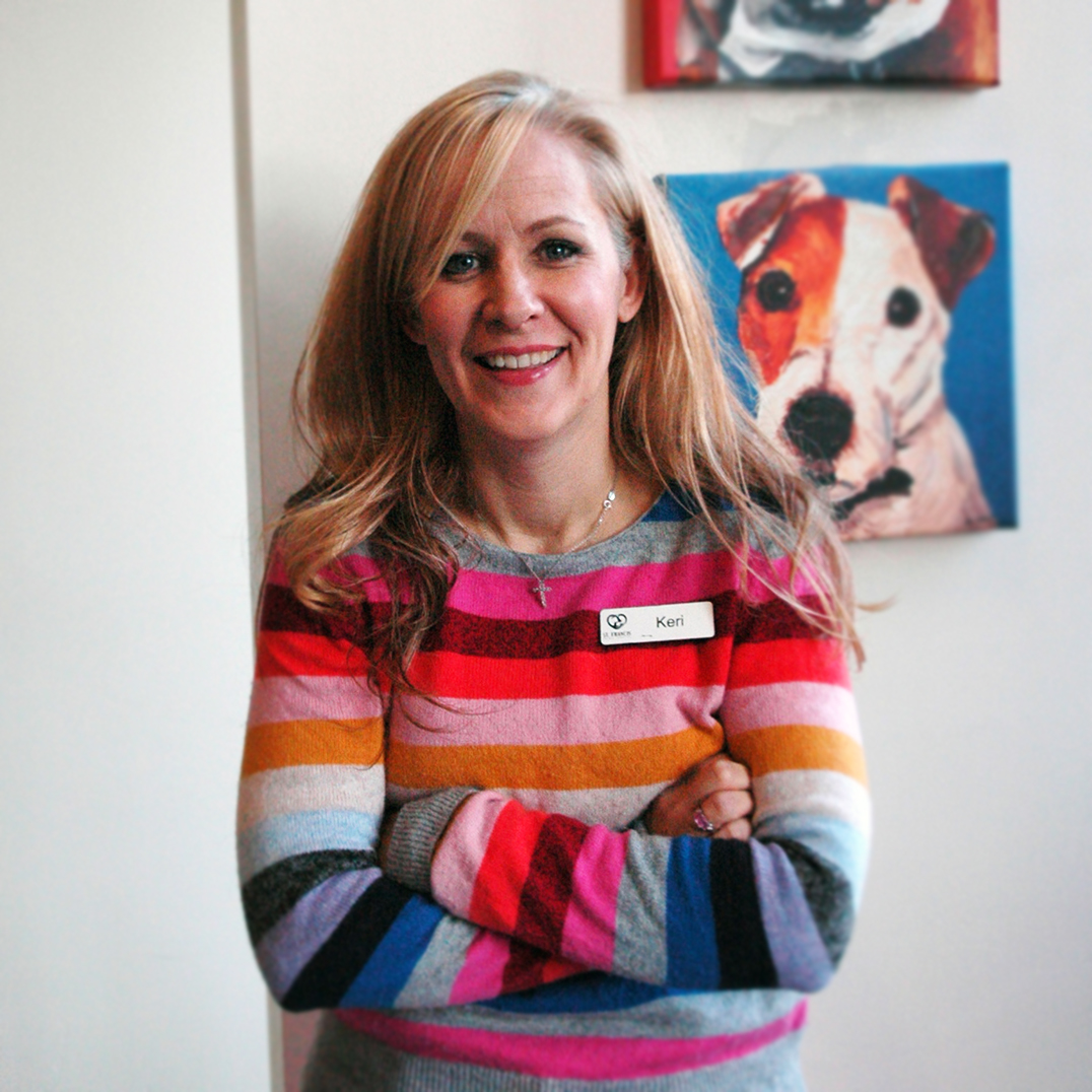 Smiling woman in a colorful striped sweater with blonde hair, standing against a white wall with a painting of a dog in the background.