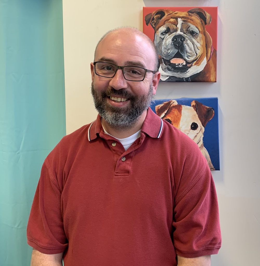 Smiling man in a red polo shirt with glasses, standing in front of a wall with two dog paintings in the background.