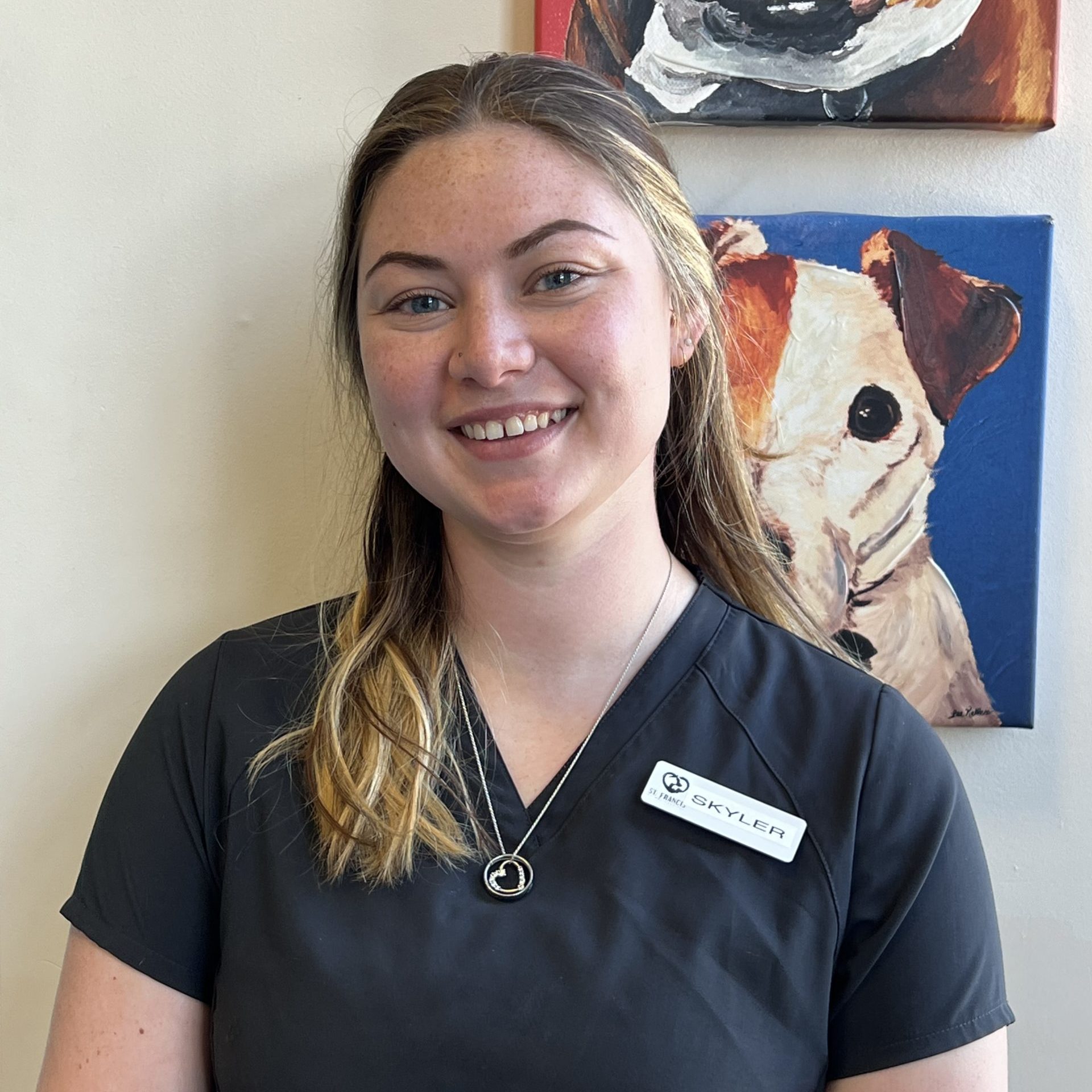 Smiling woman in a gray shirt with blonde hair, standing against a white wall with a painting of a dog in the background.