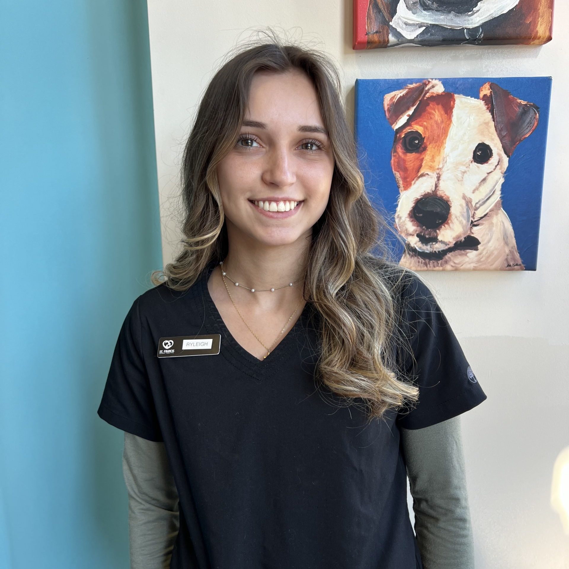 Smiling woman in a black shirt with long wavy hair, standing against a teal wall with a painting of a dog in the background.